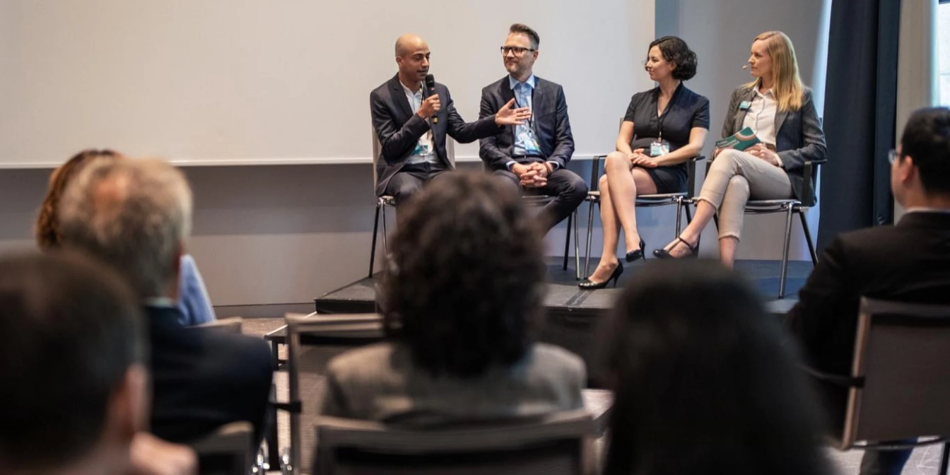A panel of four speakers seated on stage, engaging with an audience in a conference room. One speaker is holding a microphone while others listen attentively.