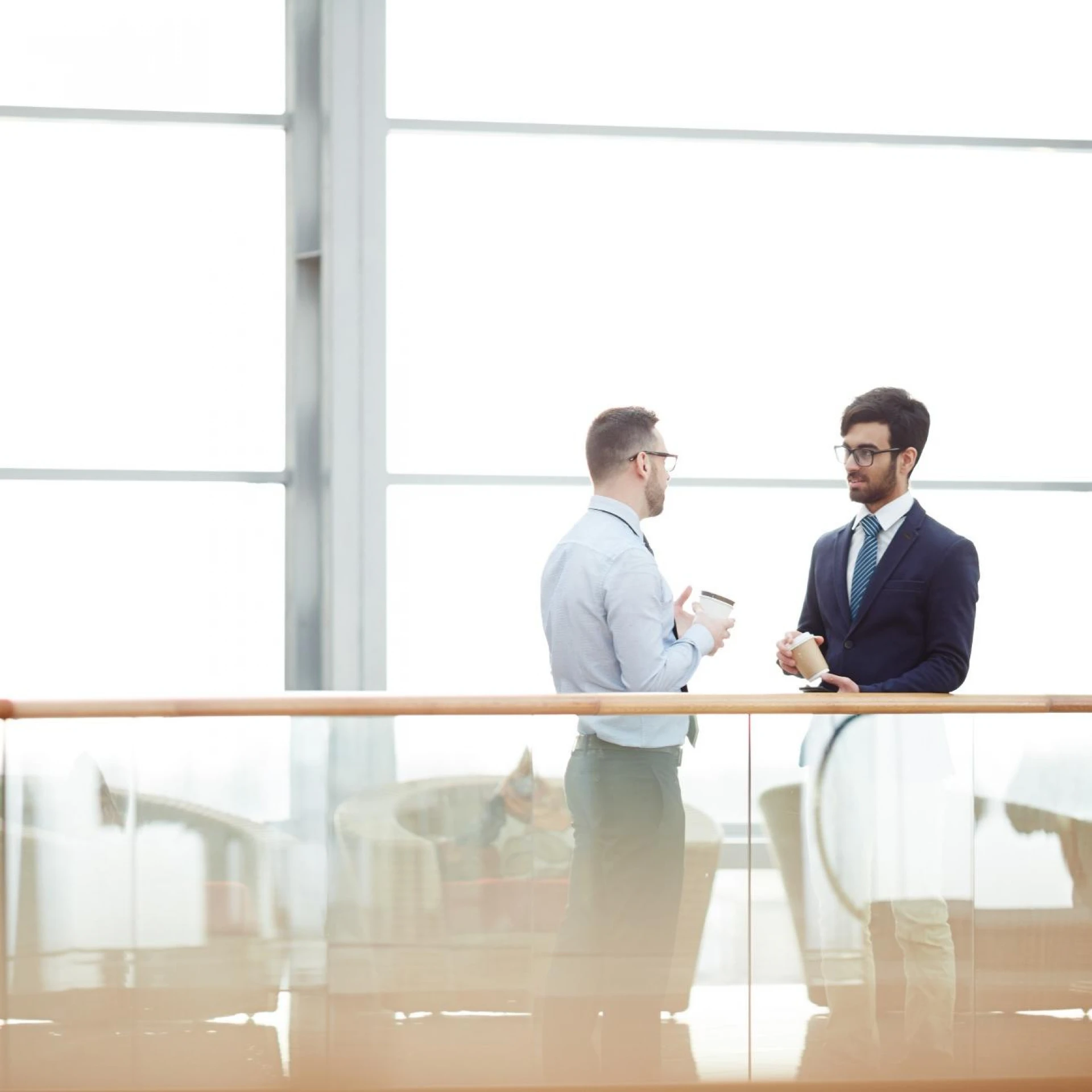 Two men in professional attire having a discussion in a bright, modern office space with large windows. Both are holding coffee cups, reflecting a casual yet professional environment.