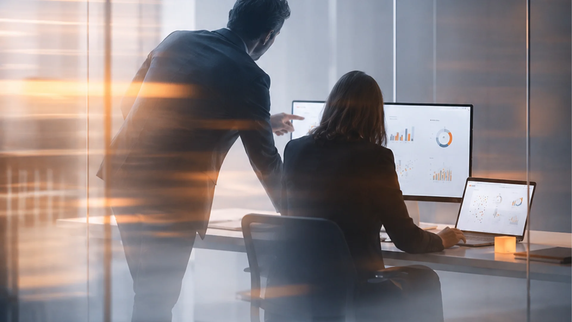 Two professionals collaborating in a modern office, one standing and pointing at a monitor displaying charts while the other is seated at a laptop, representing a different kind of partnership.